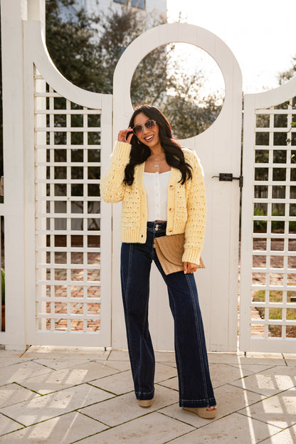 Woman wearing a yellow cardigan, white top, and blue jeans standing in front of a white lattice gate.