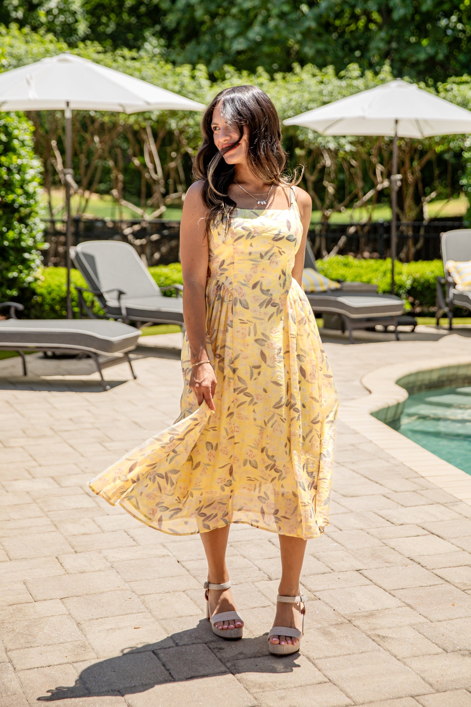 Woman in a yellow dress standing by a poolside with lounge chairs and umbrellas in the background.
