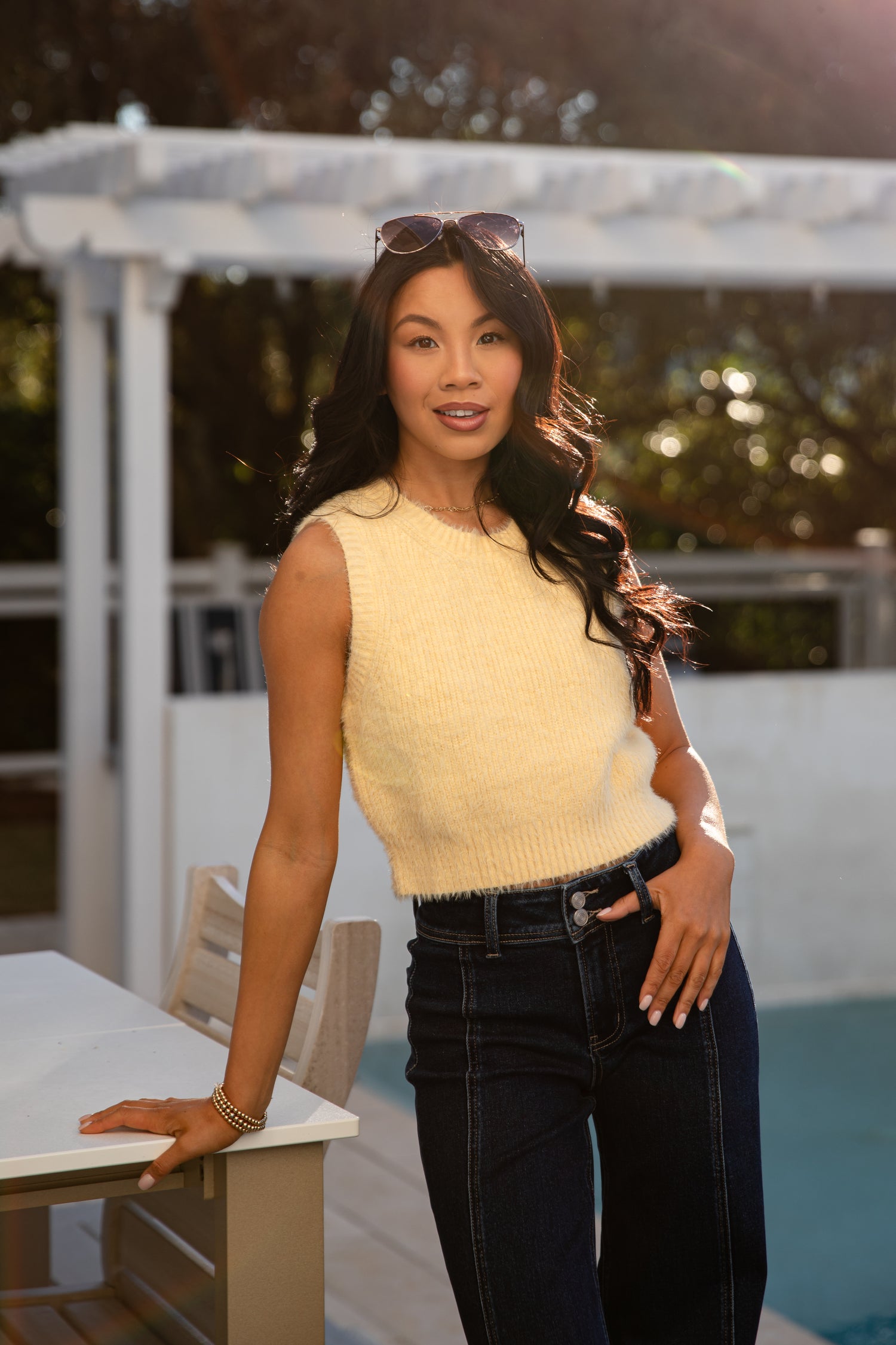 Woman in a yellow sleeveless top and dark jeans standing by a poolside.