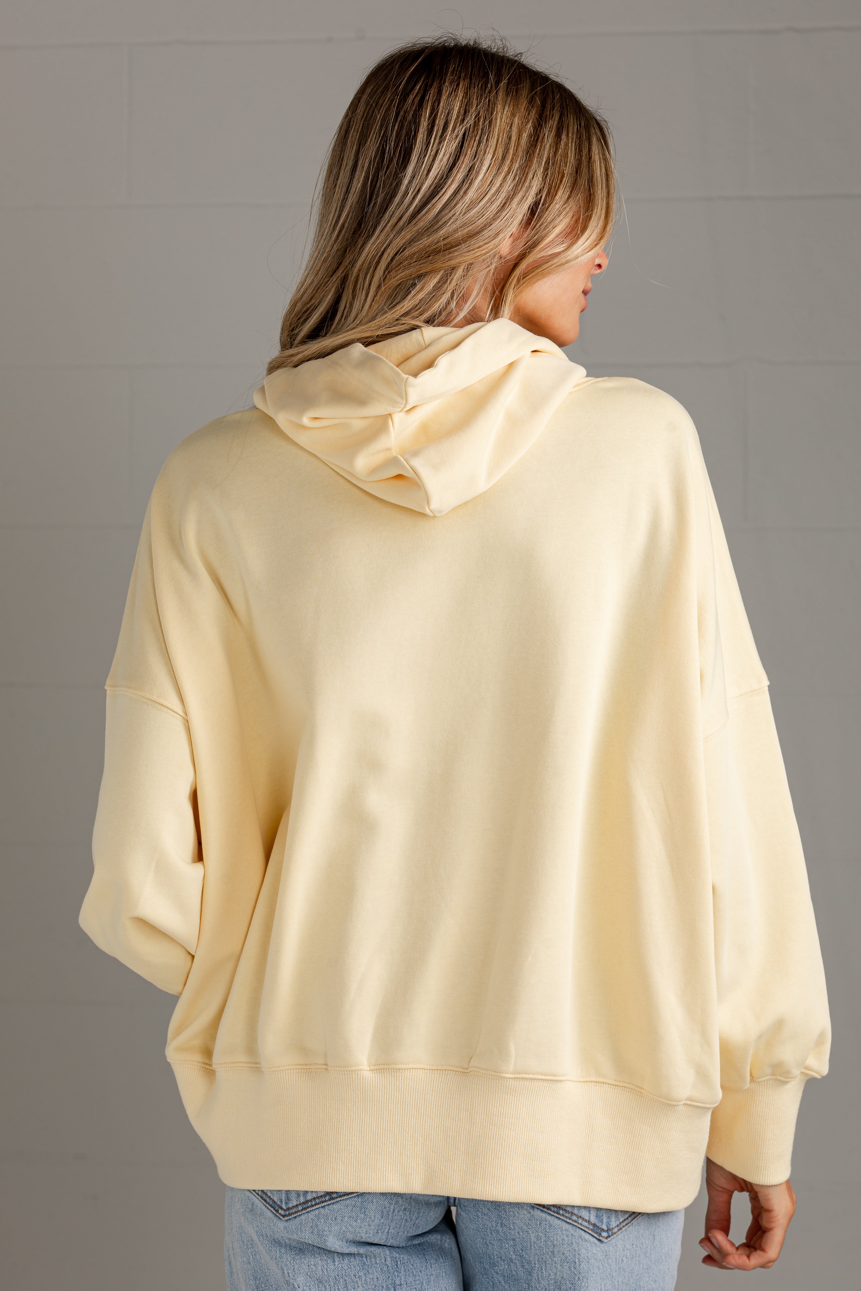 A woman viewed from the back wearing an oversized pale yellow hoodie and light blue jeans, standing against a light gray paneled wall.