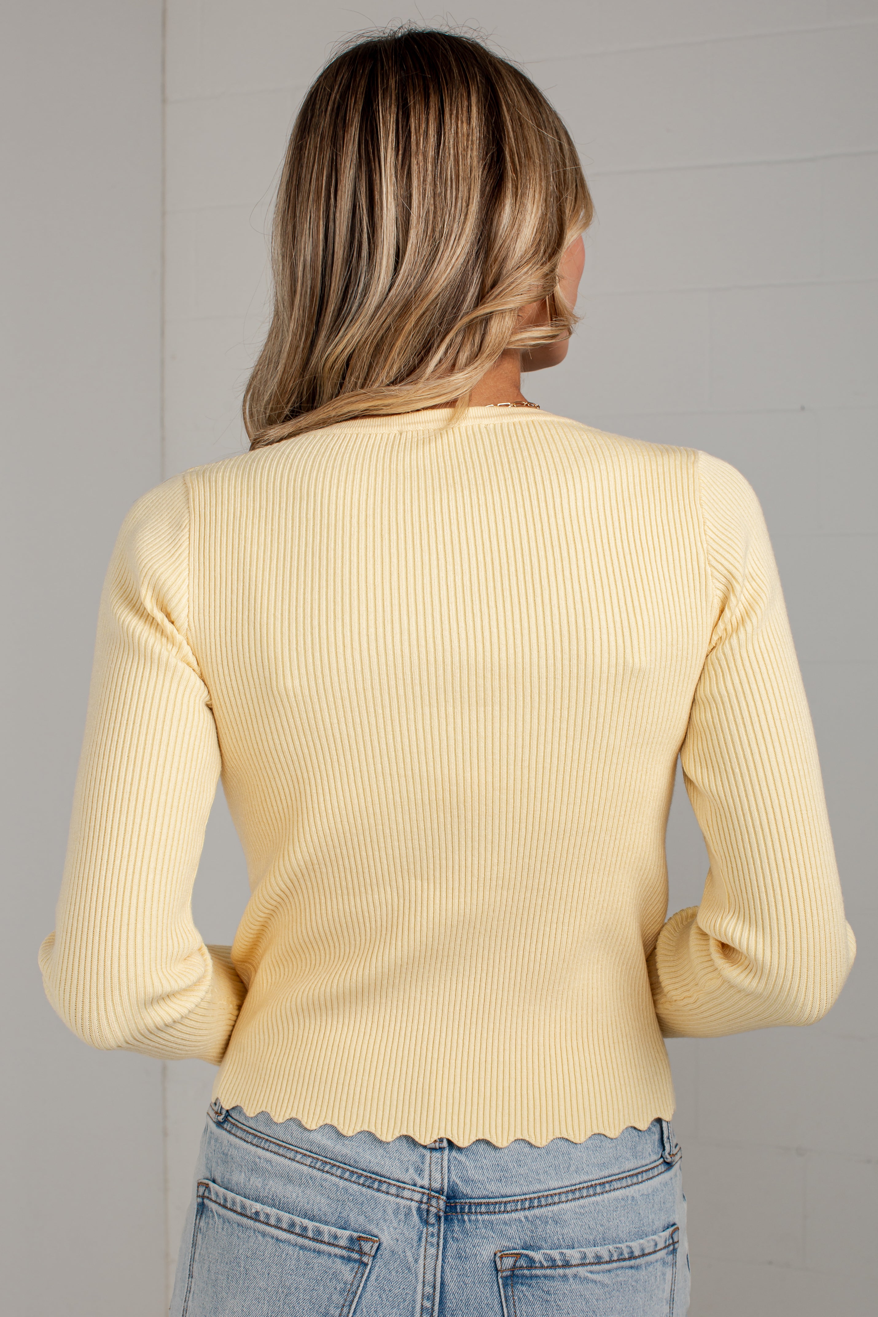 A woman viewed from behind wearing a pale yellow ribbed knit long-sleeve top with a scalloped hem and light blue jeans against a light gray wall.