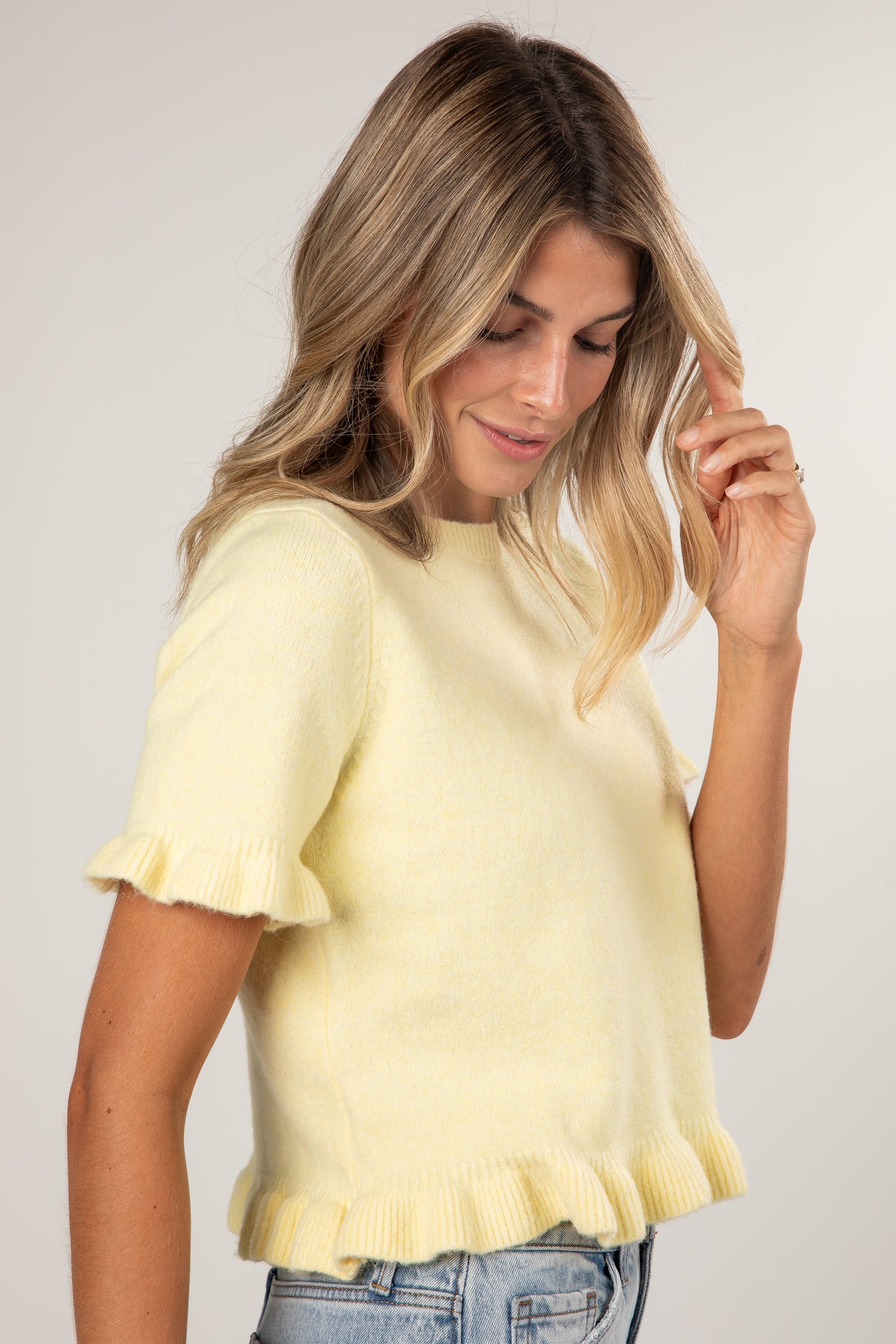 A woman wearing a pale yellow short-sleeve knit top with ruffled trim on the sleeves and hem, paired with blue jeans, photographed against a plain light gray studio background.
