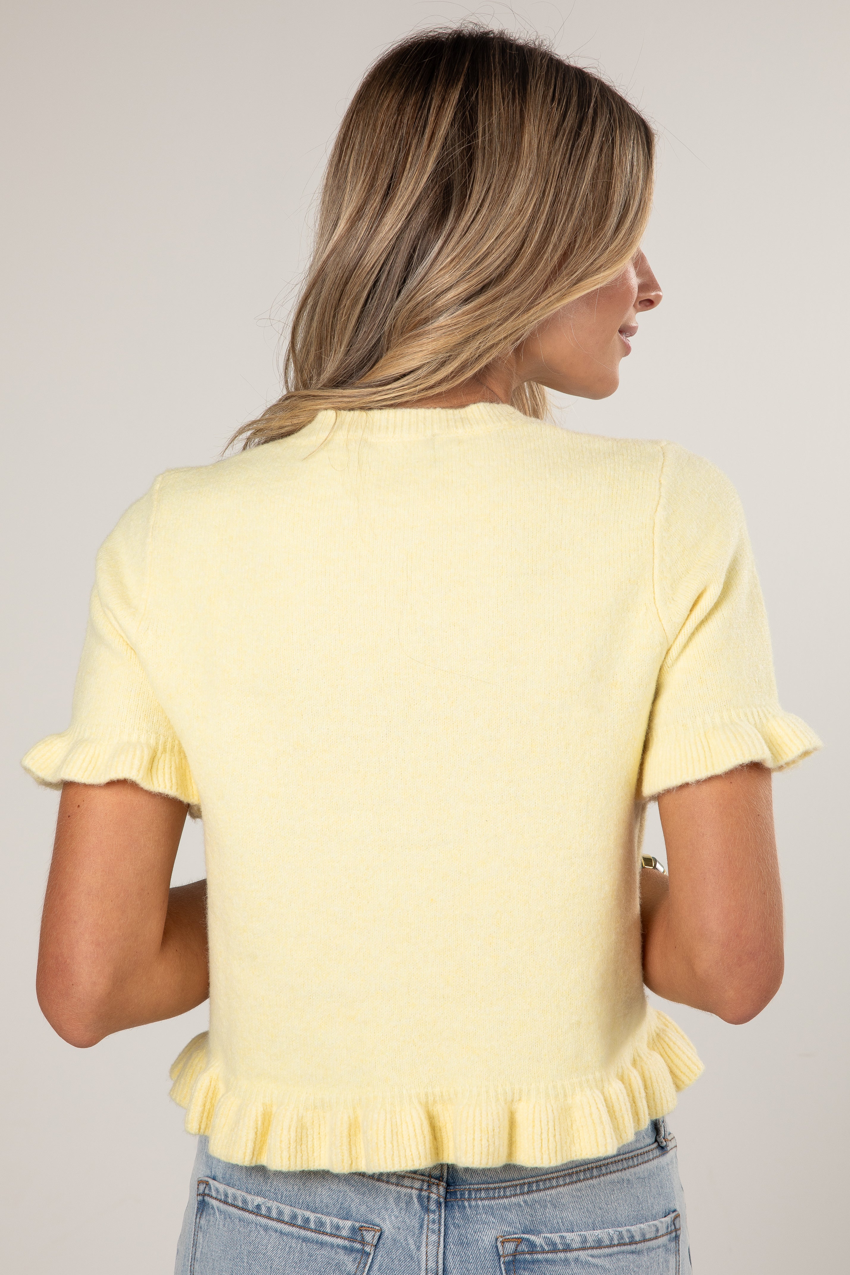 A woman shown from the back wearing a pale yellow short-sleeve knit top with ruffled trim at the sleeves and hem, paired with light blue jeans against a plain light gray studio background.