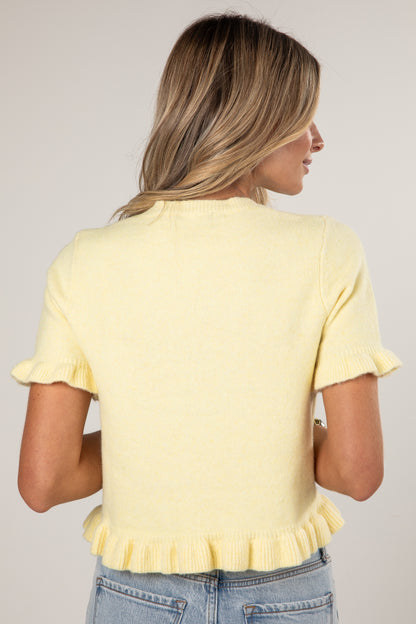 A woman shown from the back wearing a pale yellow short-sleeve knit top with ruffled trim at the sleeves and hem, paired with light blue jeans against a plain light gray studio background.