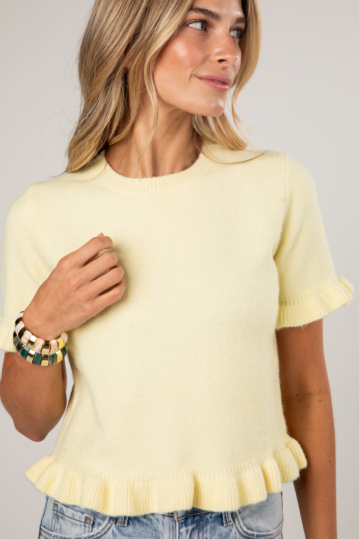 A woman wearing a pale yellow short-sleeve knit sweater with ruffled trim at the cuffs and hem, paired with blue jeans against a light gray studio background.