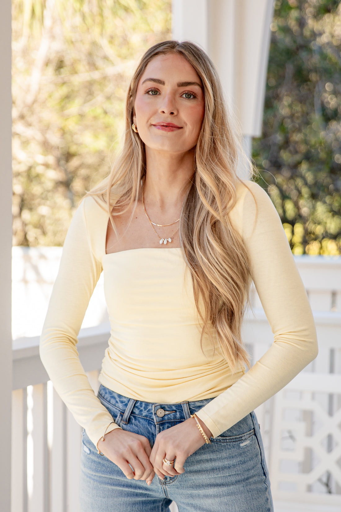 Woman wearing a yellow long-sleeve top and blue jeans on a porch.