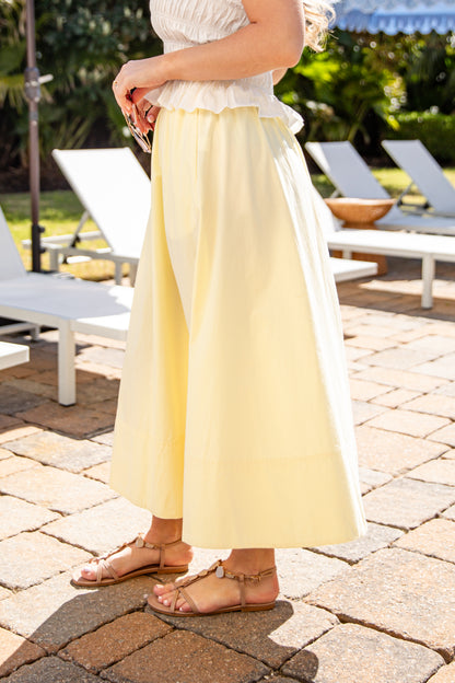 Person wearing a white top and yellow pants standing on a patio with lounge chairs and greenery in the background.