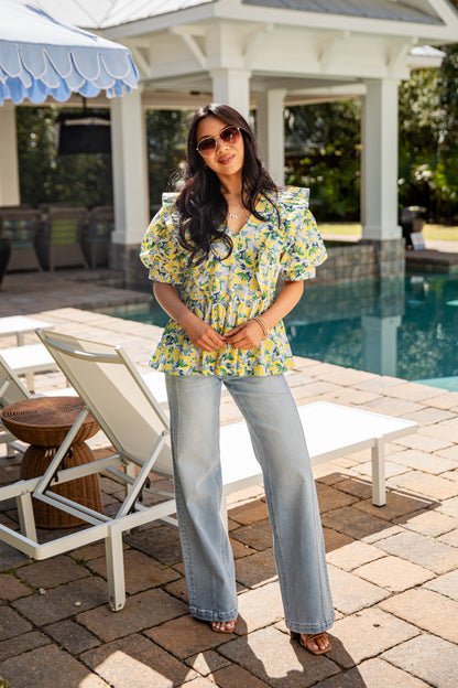 Woman in a floral top and jeans standing by a poolside with lounge chairs and a gazebo in the background.