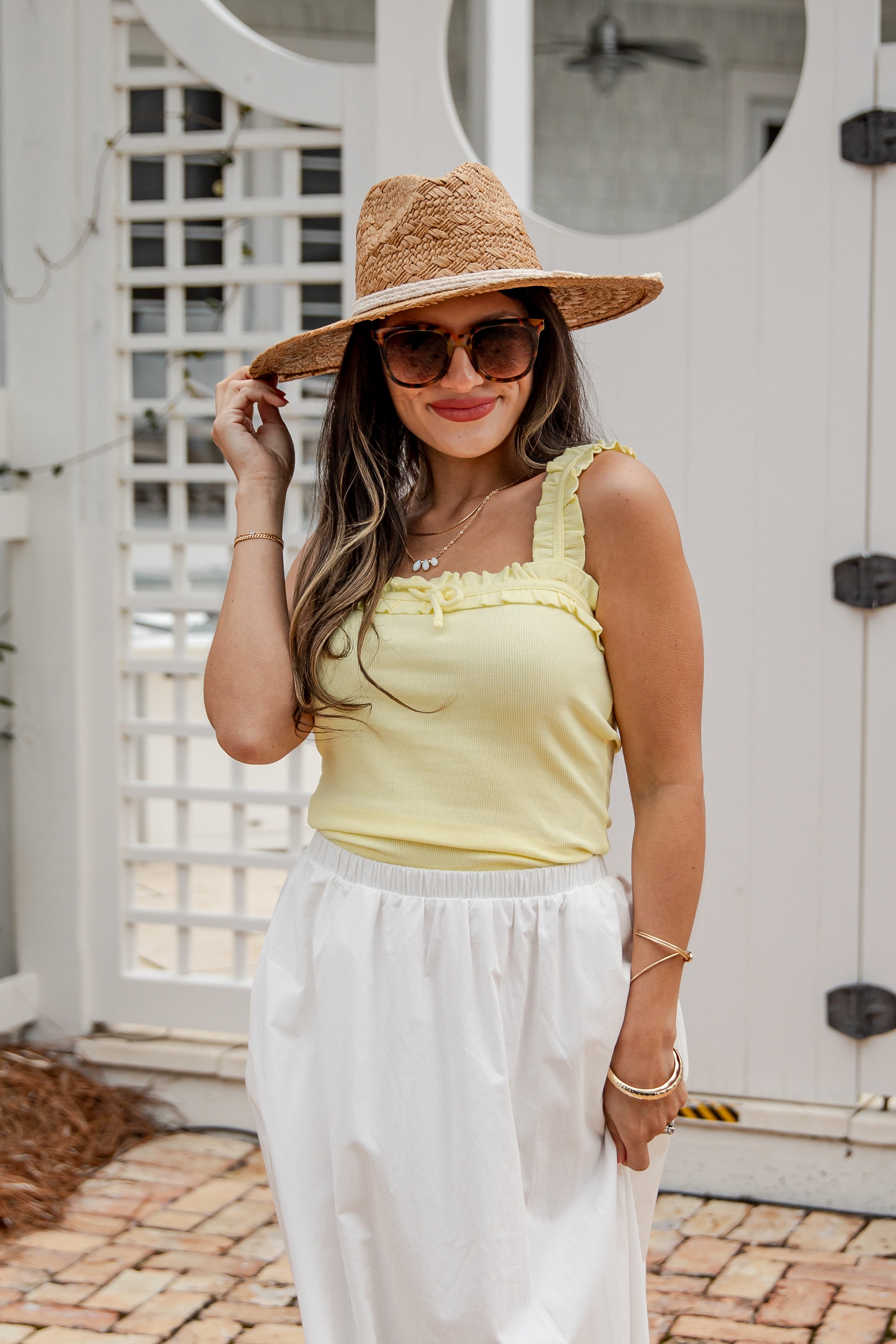 Woman wearing a yellow top and white pants with a straw hat and sunglasses, standing in front of a white door.