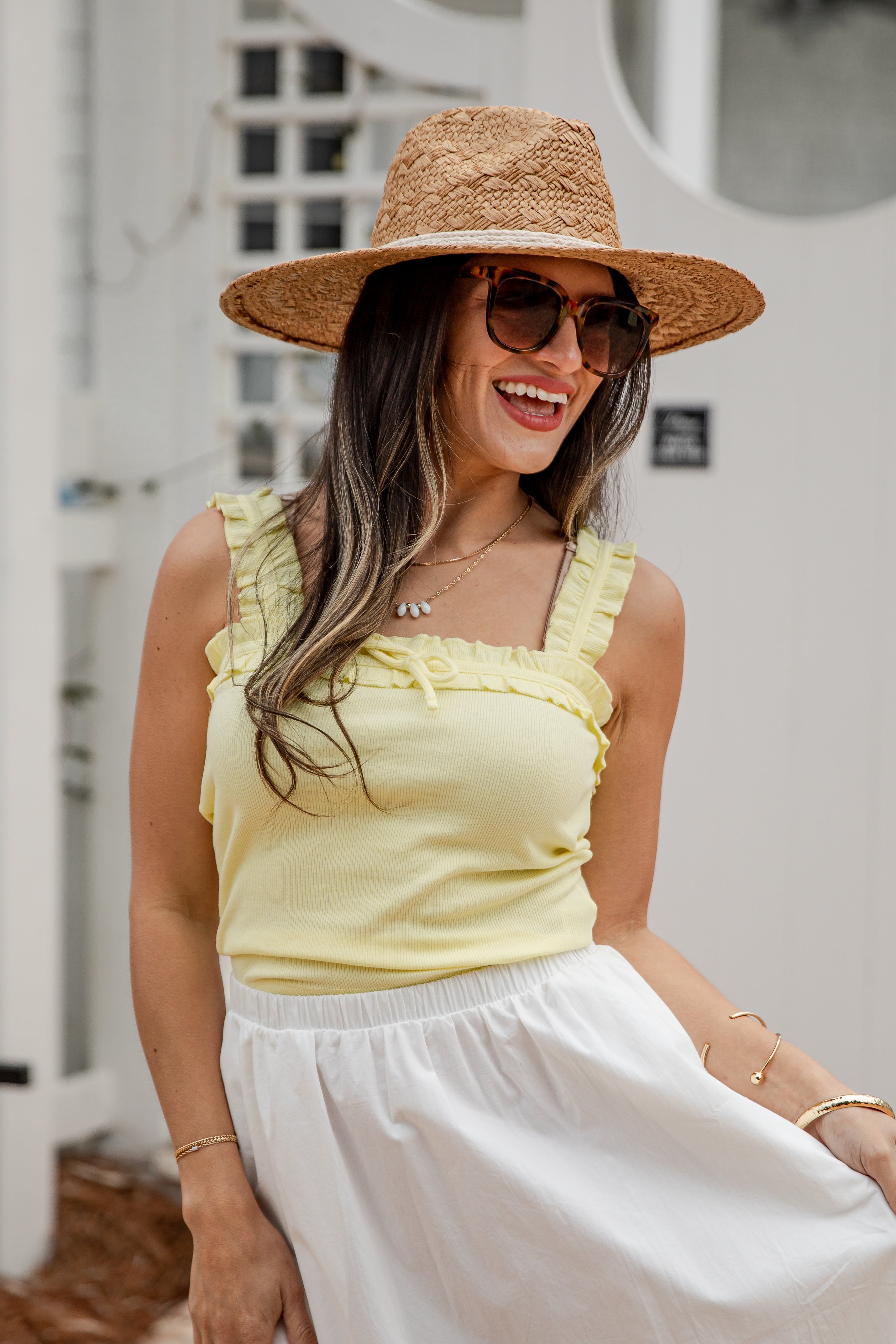 Woman wearing a yellow top and white skirt with a straw hat and sunglasses, standing against a blurred outdoor background.