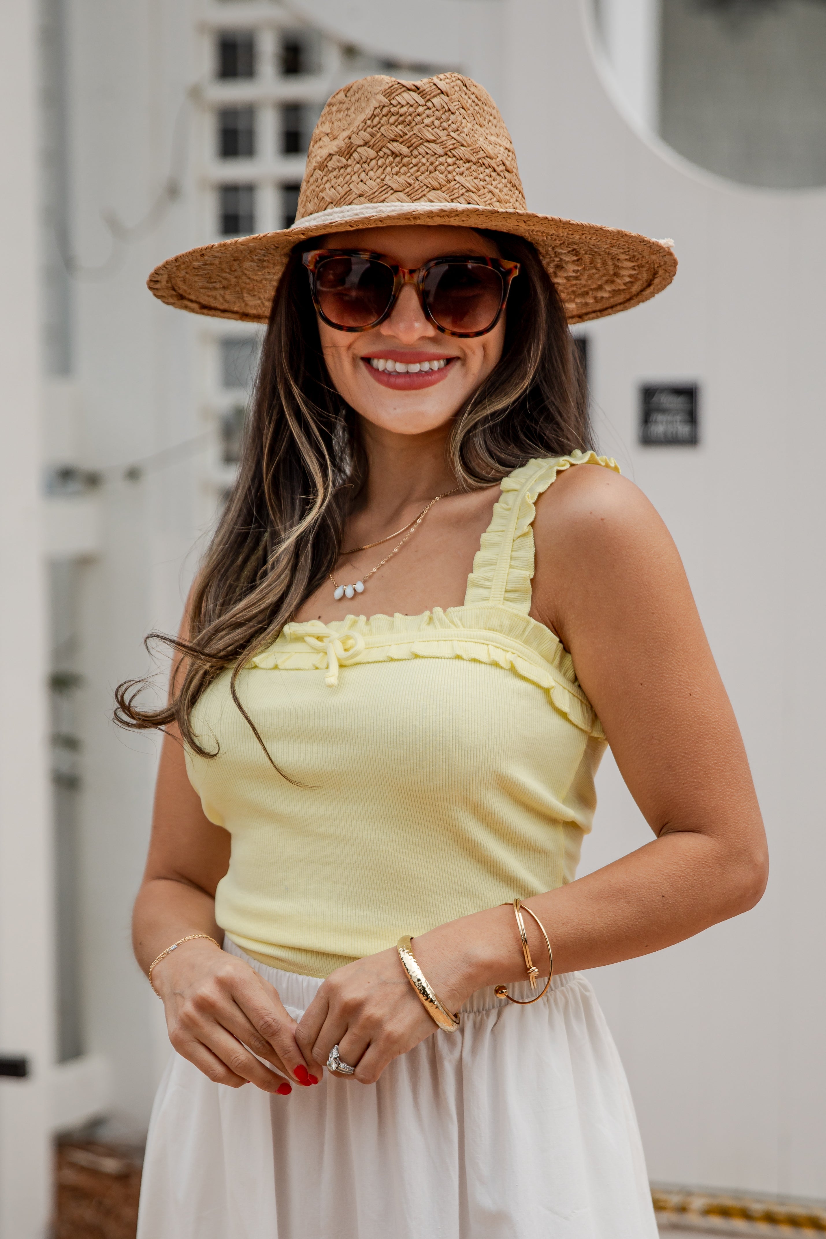 Woman wearing a yellow top and straw hat with sunglasses, standing outdoors.