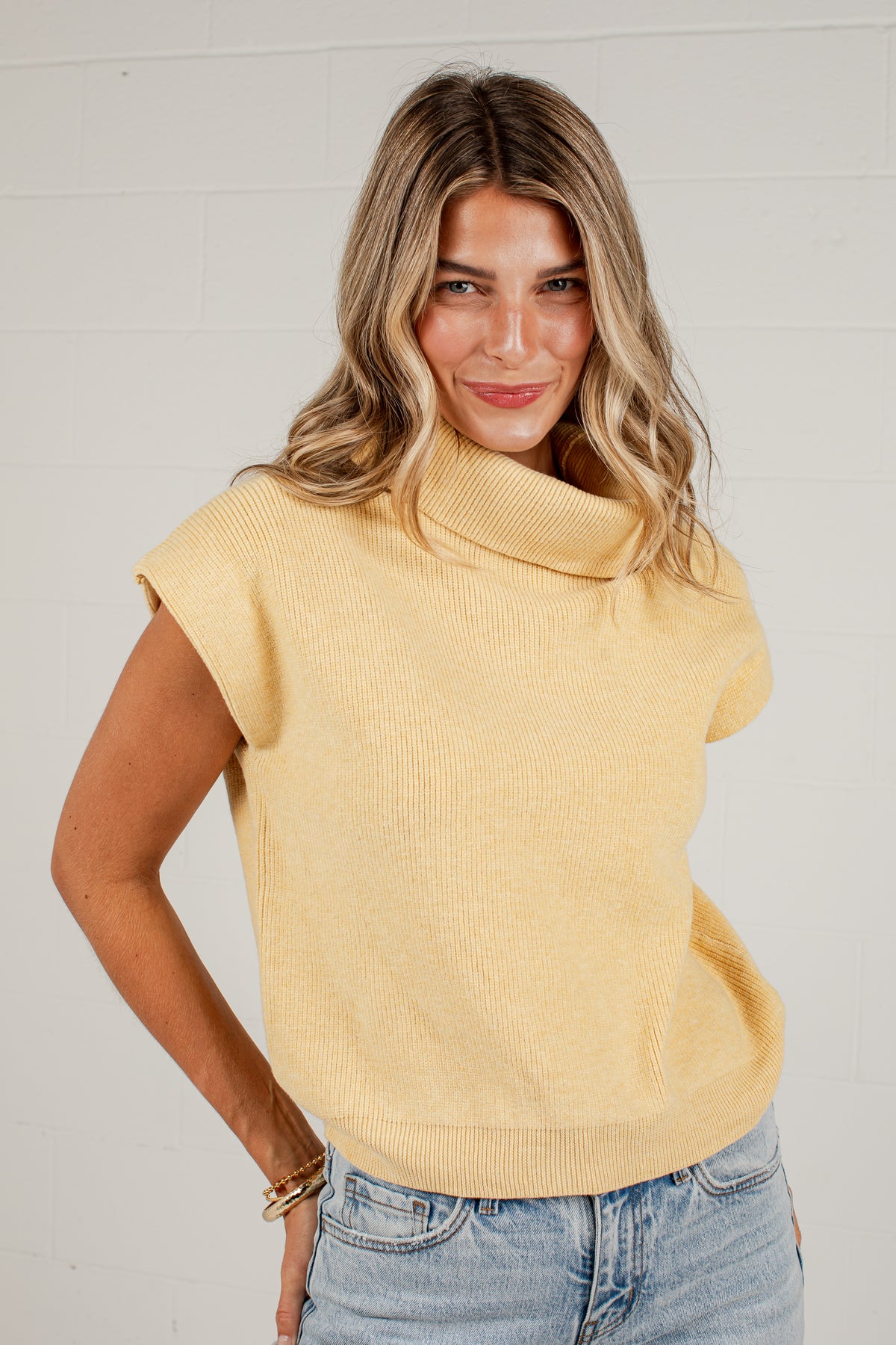 A woman wearing a pale yellow ribbed knit cowl-neck sweater with cap sleeves and light-wash blue jeans, standing against a white brick wall.