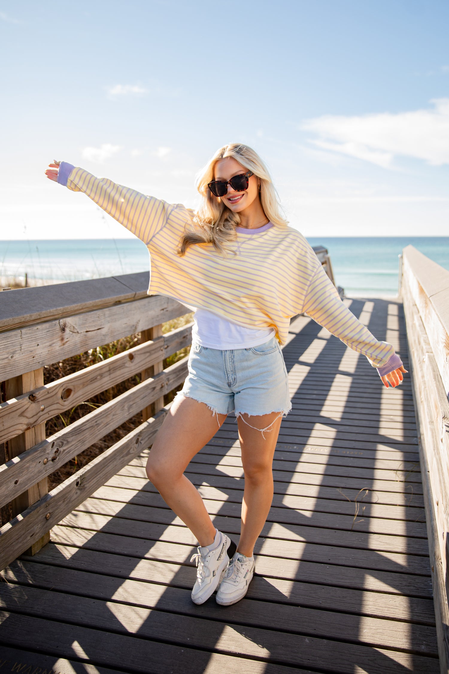 Woman standing on a wooden boardwalk by the beach, wearing a striped sweater and denim shorts.