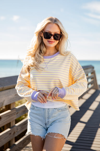 Woman wearing a yellow and white striped shirt with blue shorts on a wooden deck by the ocean.