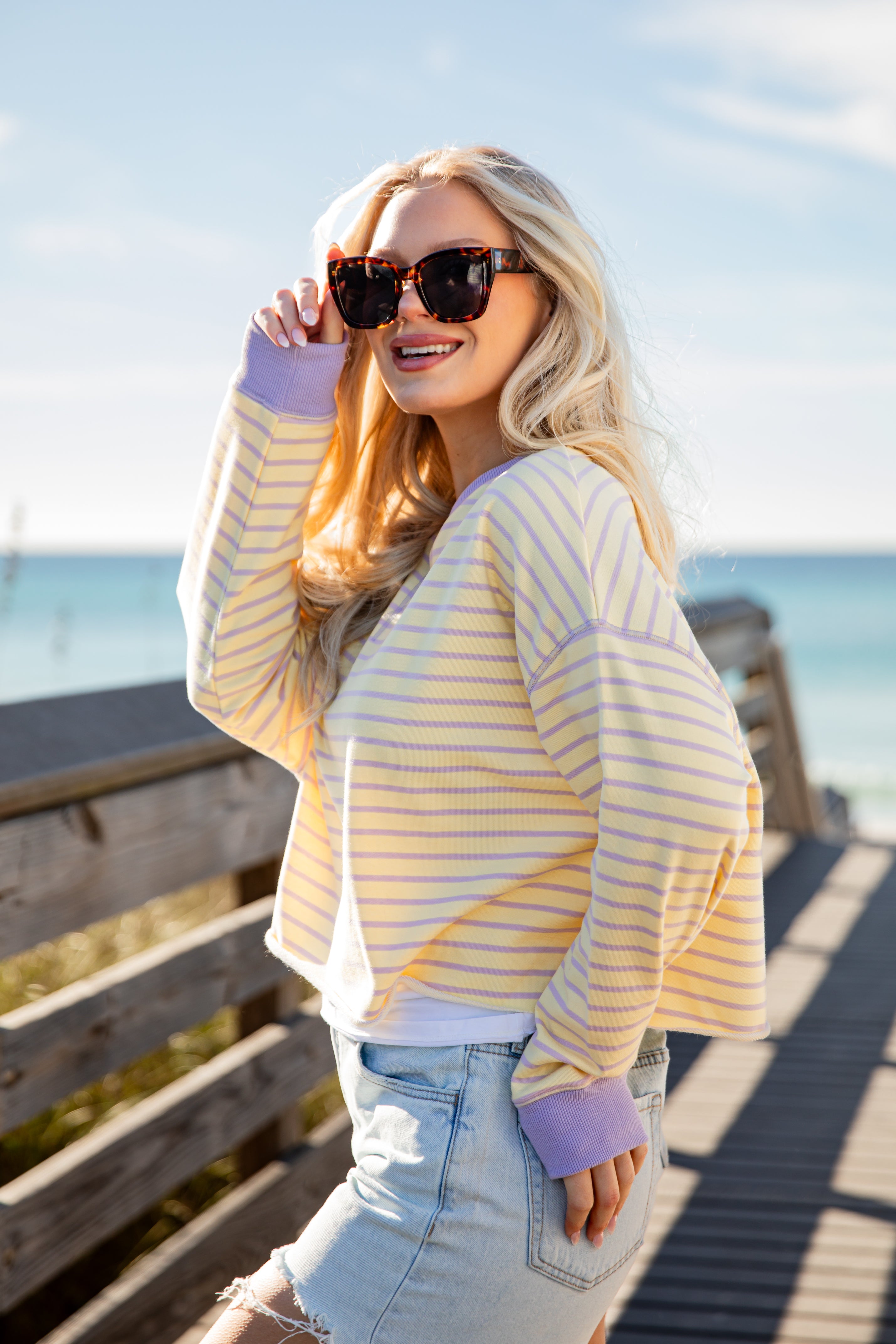 Woman wearing a yellow and white striped sweater and sunglasses by the ocean.