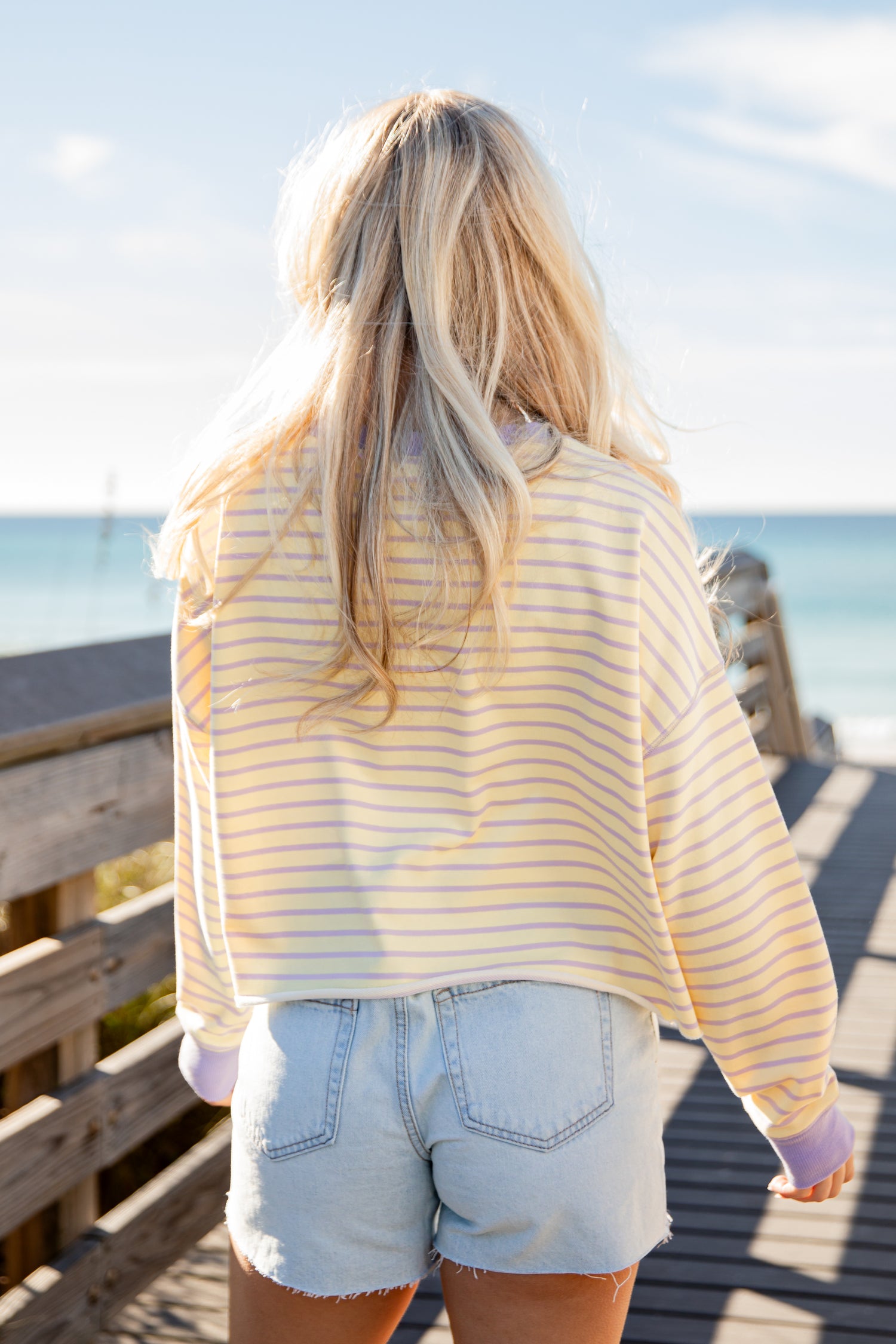 Person wearing a yellow striped shirt and denim shorts standing on a wooden pier with ocean view.
