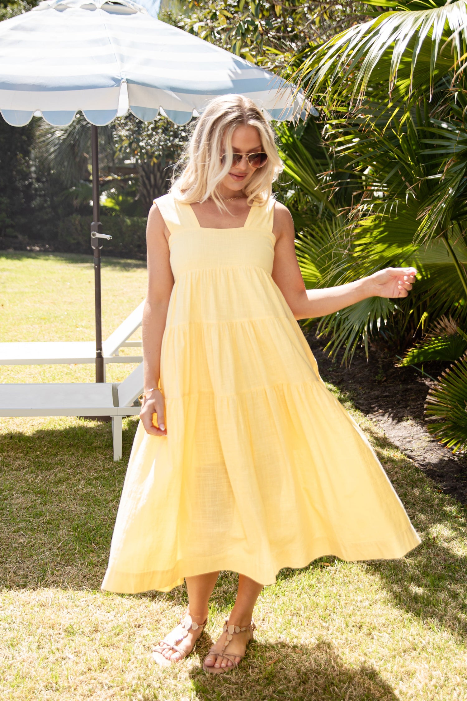 Woman in a yellow dress standing outdoors under a white umbrella with greenery around.