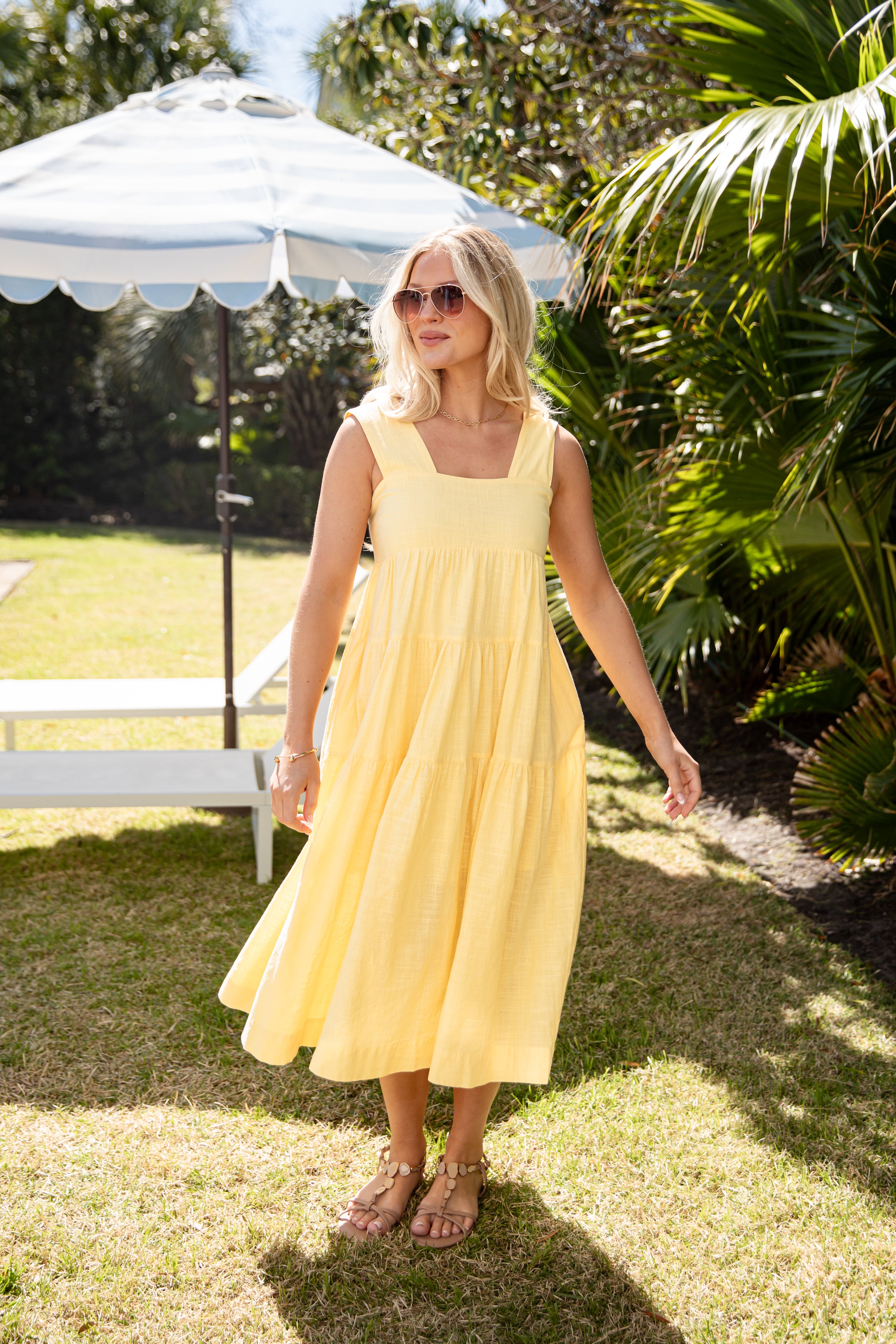 Woman in a yellow dress standing outdoors under an umbrella with greenery around.