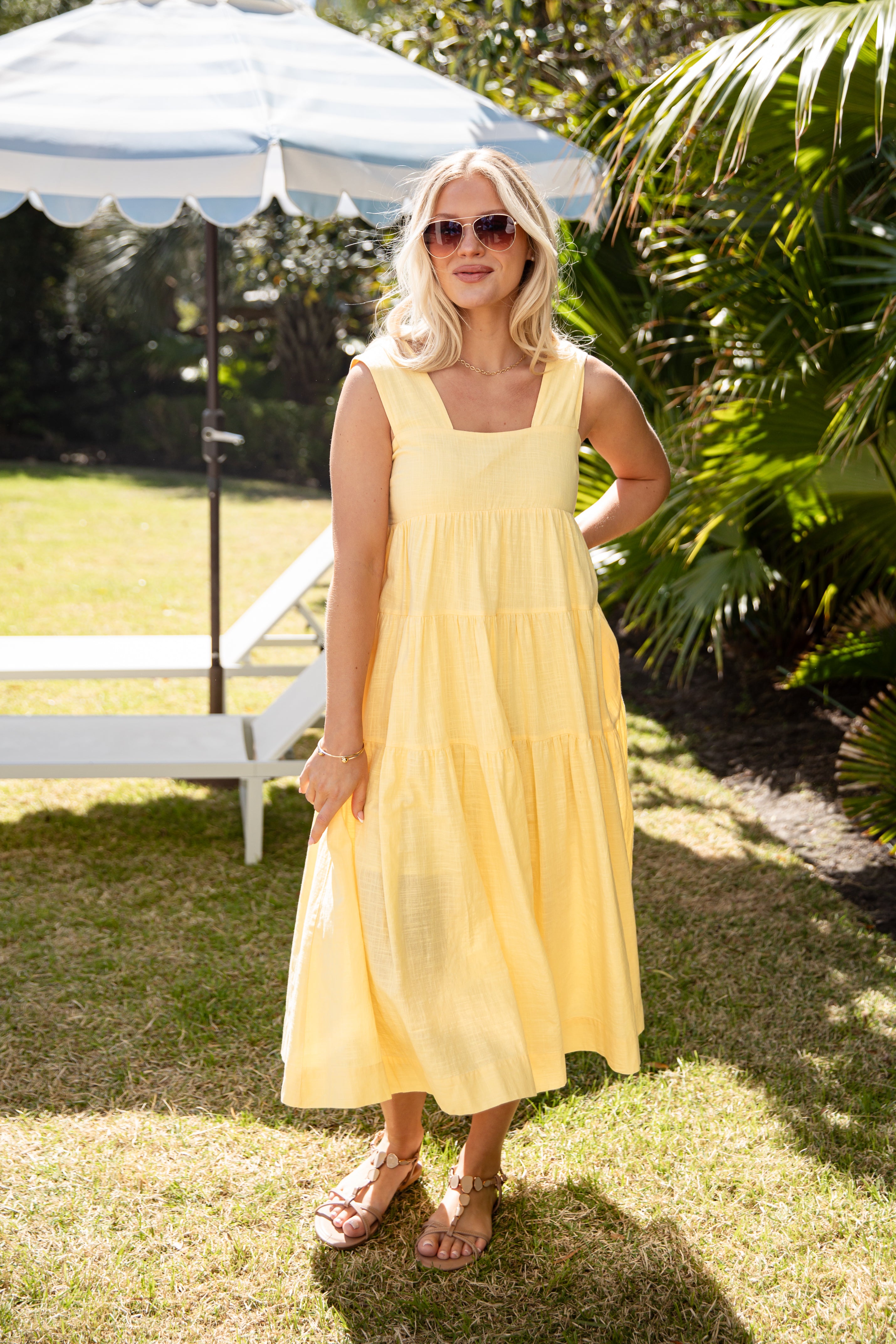 Woman in a yellow dress standing outdoors under a white umbrella with greenery in the background.