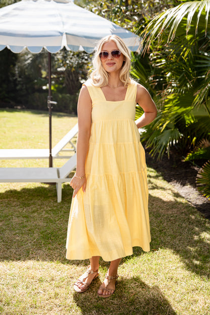 Woman in a yellow dress standing outdoors under a white umbrella with greenery in the background.
